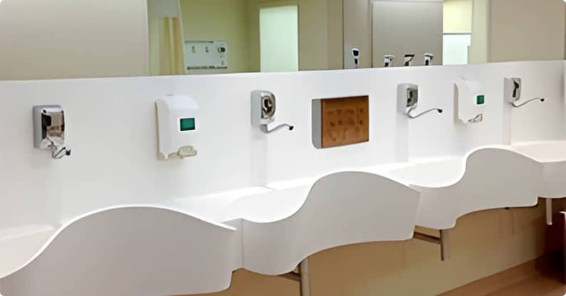 Móveis planejados em um conjunto de pias brancas modernas e curvadas em um ambiente hospitalar, com dispensadores de sabão e torneiras com sensores fixados em uma parede branca