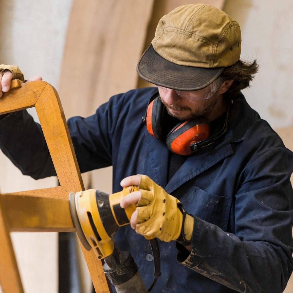 Um homem, vestindo uma camisa de manga comprida azul, boné marrom e luvas de proteção, usa uma lixadeira elétrica para lixar a perna de uma cadeira de madeira em sua oficina